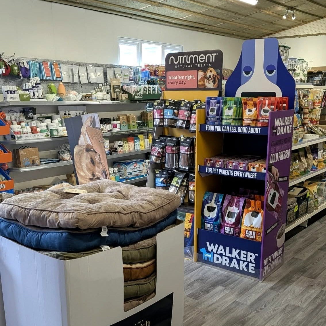 A retail store aisle showing pet bedding products, including a large plush dog bed on a white display stand in the foreground and a purple Walker Drake pet food display shelf with dog treats.
